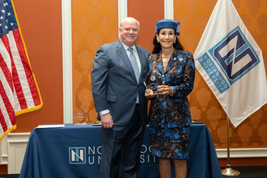 A man in a suit and a woman in a blue outfit pose with an award in front of a Northwood University automotive table, flag, and an American flag.