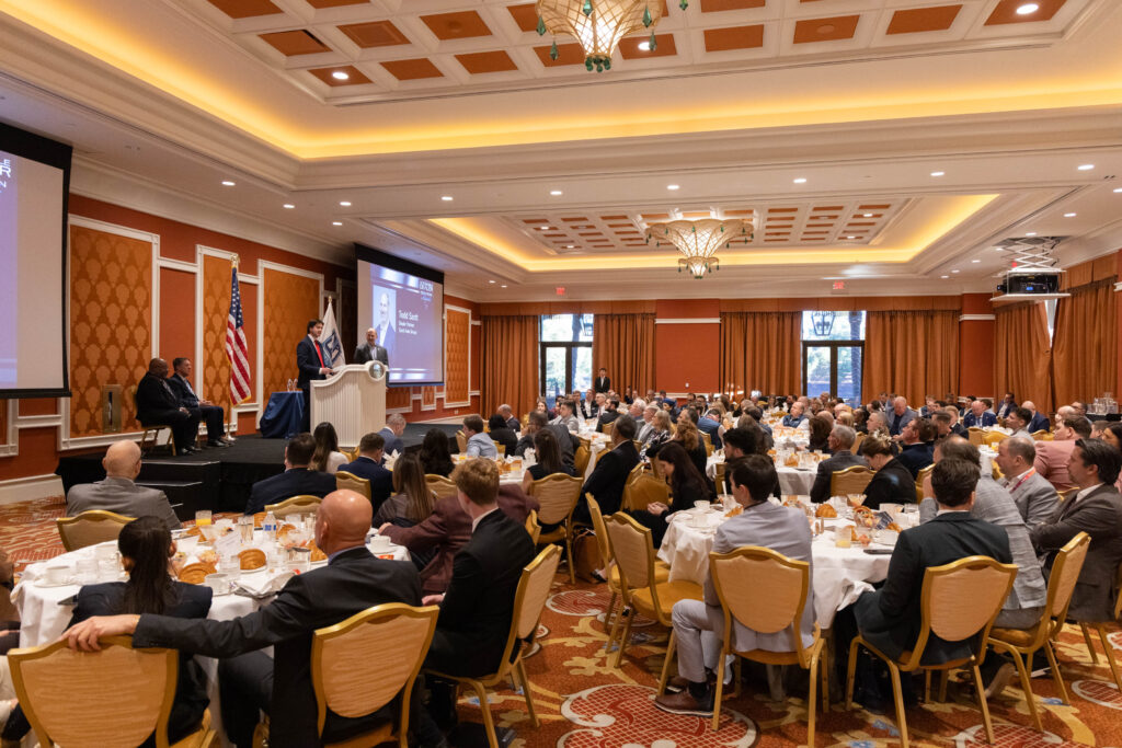 A large group of people seated at round tables in a university banquet hall listen to speakers at a podium during a formal automotive event.