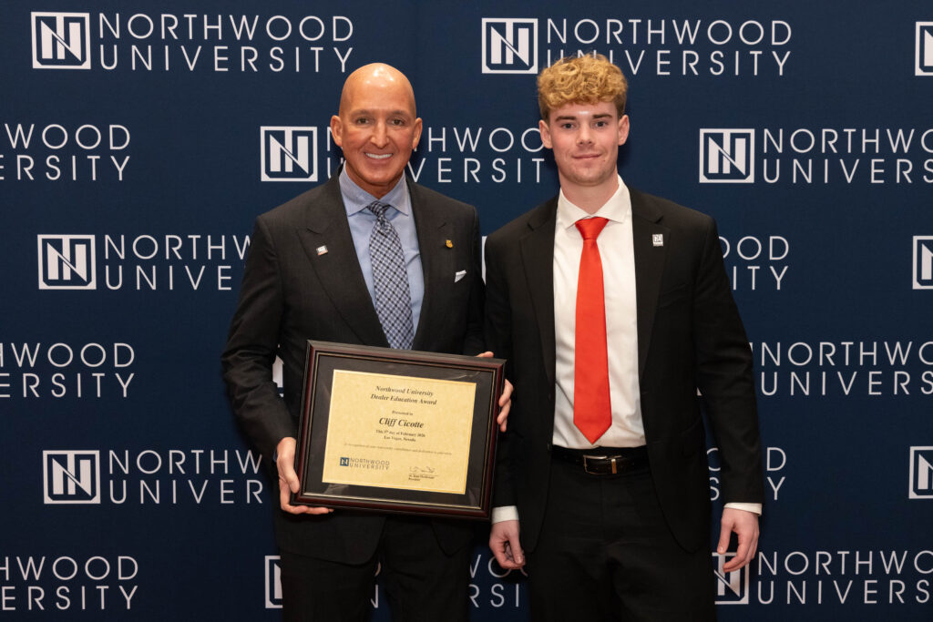 Two men in business attire stand in front of a Northwood University backdrop; one holds a framed certificate that reads "Cliff Coone," highlighting university automotive achievements.