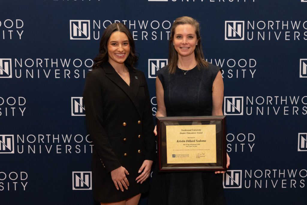 Two women stand in front of a Northwood University automotive backdrop; one holds a framed certificate while both smile at the camera.