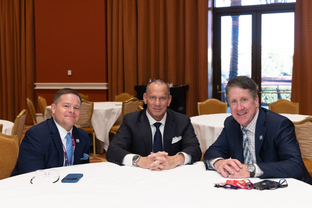 Three men in suits sit at a round table in a university conference room, with automotive documents spread before them, brown curtains, and empty chairs in the background.