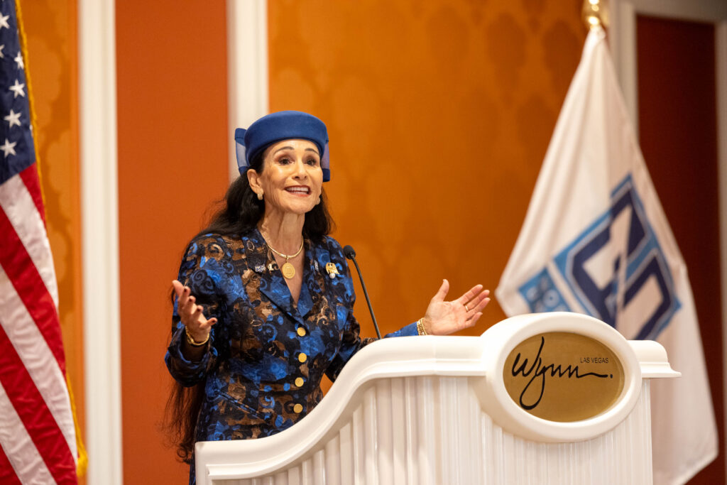 A woman in a blue patterned suit and hat speaks at a podium with the Wynn logo, addressing university and automotive leaders, with an American flag and another flag in the background.
