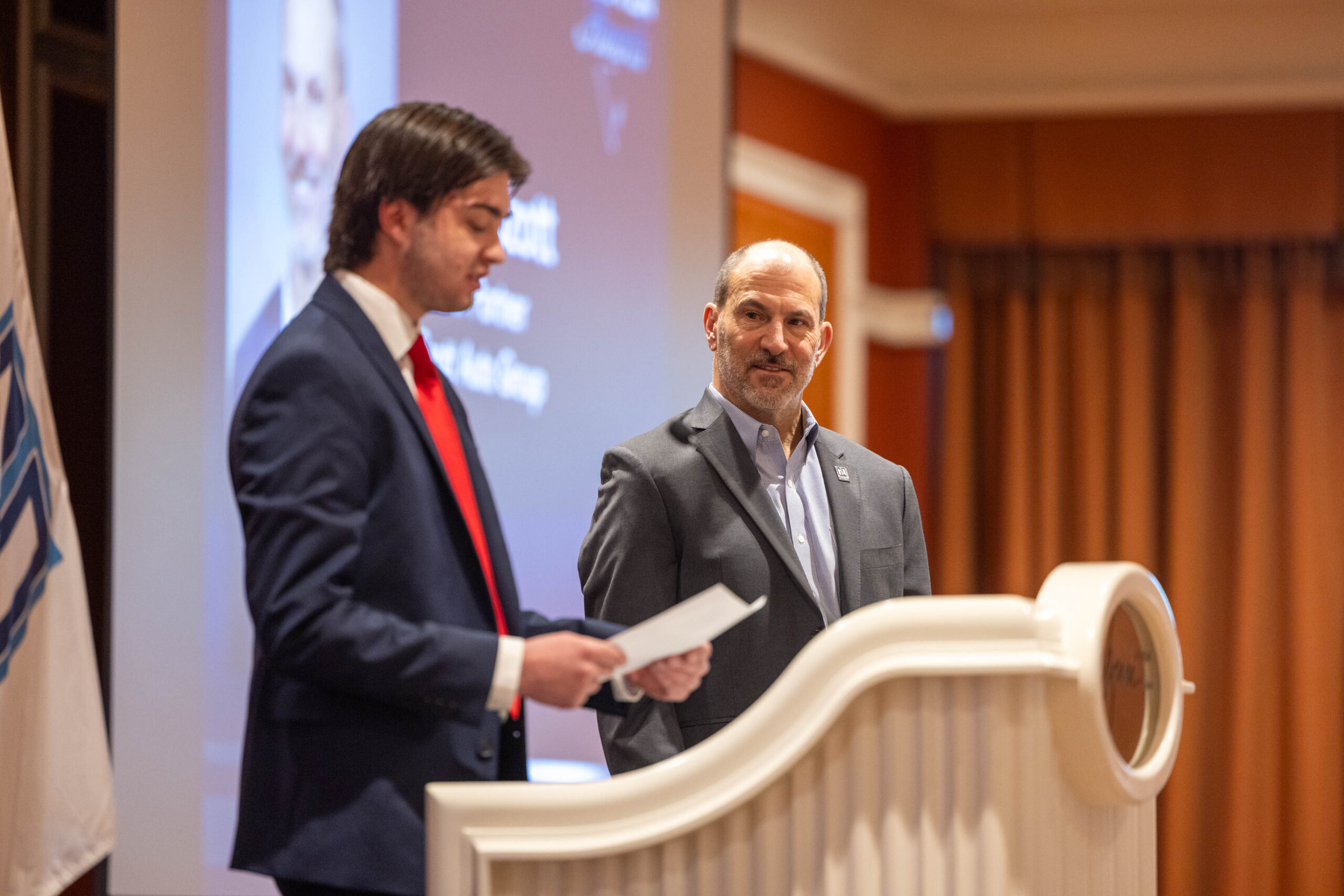 Two men in suits stand at a podium during a formal university event, with a presentation screen showcasing automotive topics and a flag visible in the background.