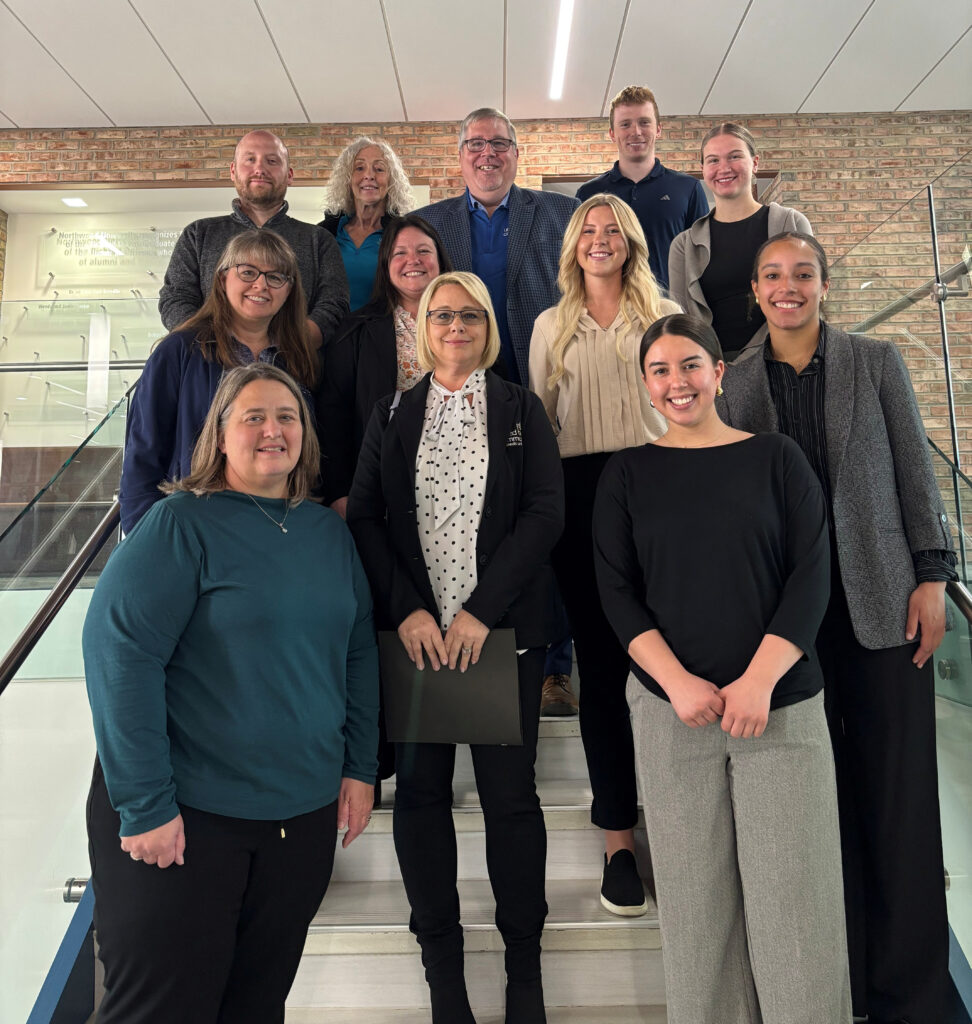 A group of twelve DeVos Students poses together on an indoor staircase, smiling toward the camera in a professional setting within the Great Lakes Bay Region.
