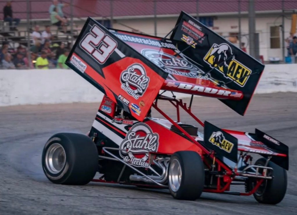 A red and black sprint car with the number 23 and various sponsor logos races on a dirt track, showcasing Charlie Baur’s motorsports career as spectators watch from behind a fence.