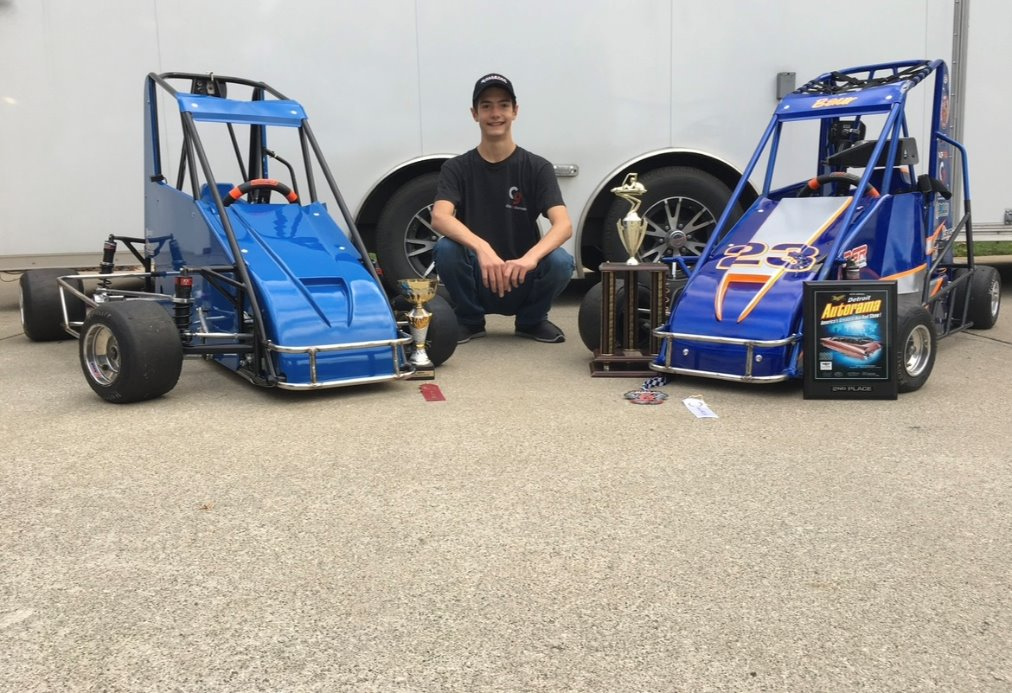 A person kneels on concrete between two small blue race cars—three trophies and a framed certificate in front—celebrating Charlie Baur’s early motorsports career with visible business discipline and pride.
