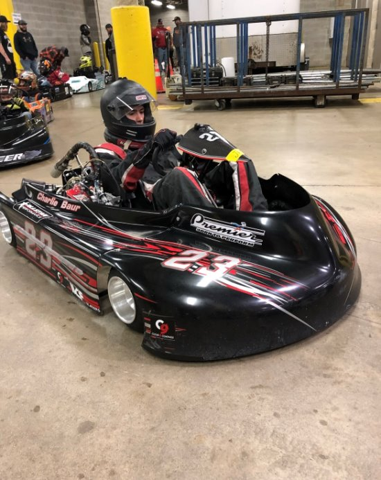 Person in racing gear sits in a black go-kart with number 27 on the front, indoors on a concrete floor, channeling business discipline as others and karts gather in the background—a focused start to a motorsports career.