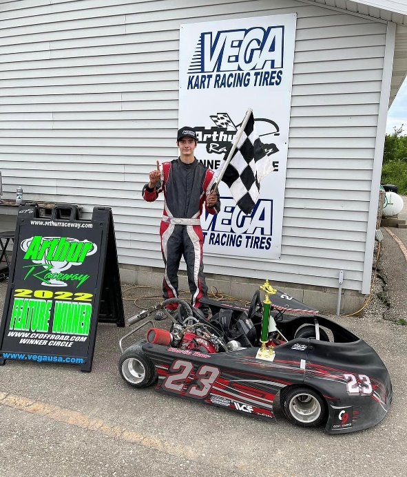 A person in a racing suit stands next to a go-kart holding a checkered flag, in front of a sign for kart racing tires and a podium board showing "Feature Winner," embodying the business discipline essential for a successful motorsports career.