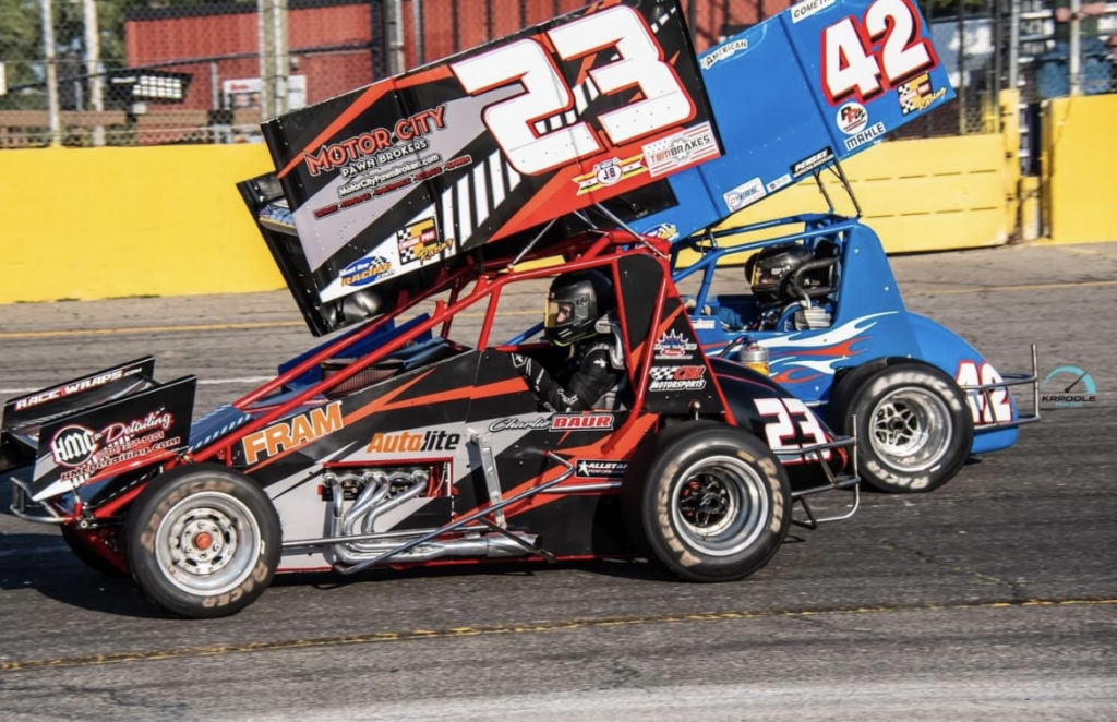 Two sprint cars, numbered 23 and 42, race side by side on an asphalt track beside a yellow wall, embodying the business discipline that has defined Charlie Baur’s motorsports career.