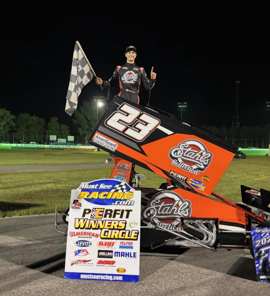 Race car driver Charlie Baur stands on car number 23 holding a checkered flag, next to a winners' circle sign on a racetrack at night, celebrating another milestone in his impressive motorsports career.