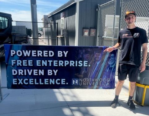 A person stands next to a Northwood University banner that reads, "Powered by Free Enterprise. Driven by Excellence," outside near a building and fence.