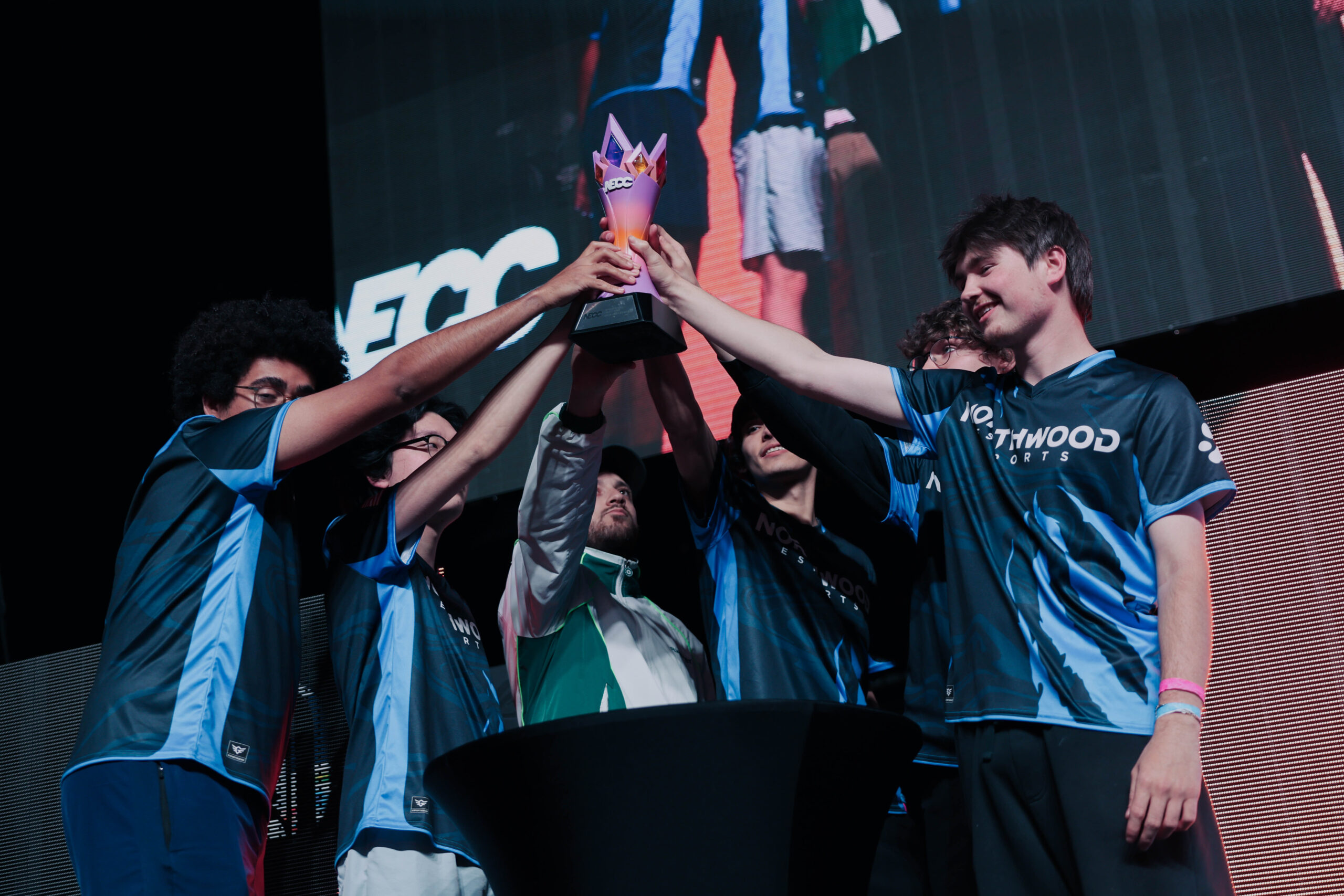 A group of esports players wearing team jerseys lift a trophy together on stage, with a large screen behind them displaying their image.