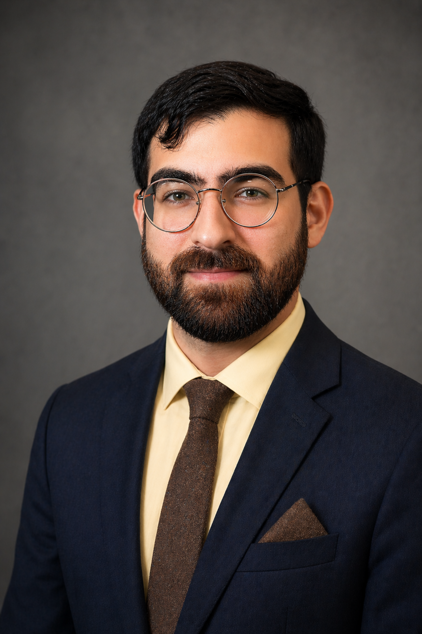 A man with dark hair, a beard, and glasses, wearing a navy suit, yellow shirt, and brown tie, poses against a plain gray background.