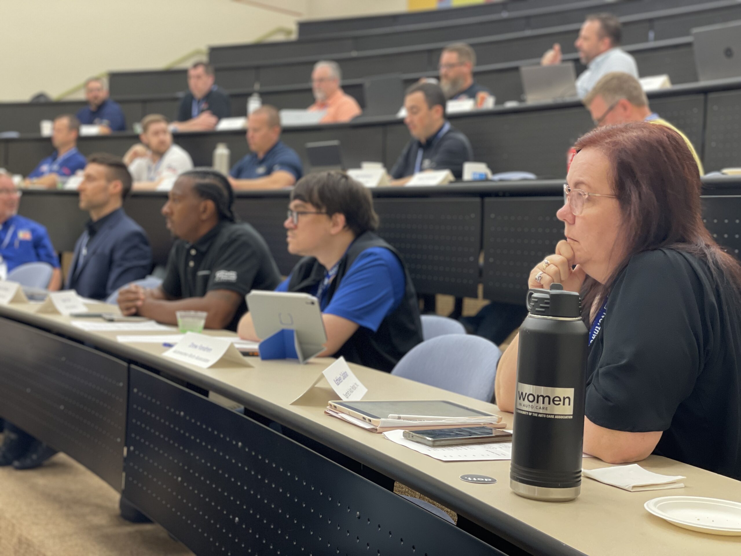 People sit in rows of a lecture hall, listening attentively with laptops and notepads on their desks. A woman in the front has a large water bottle labeled “women.”.