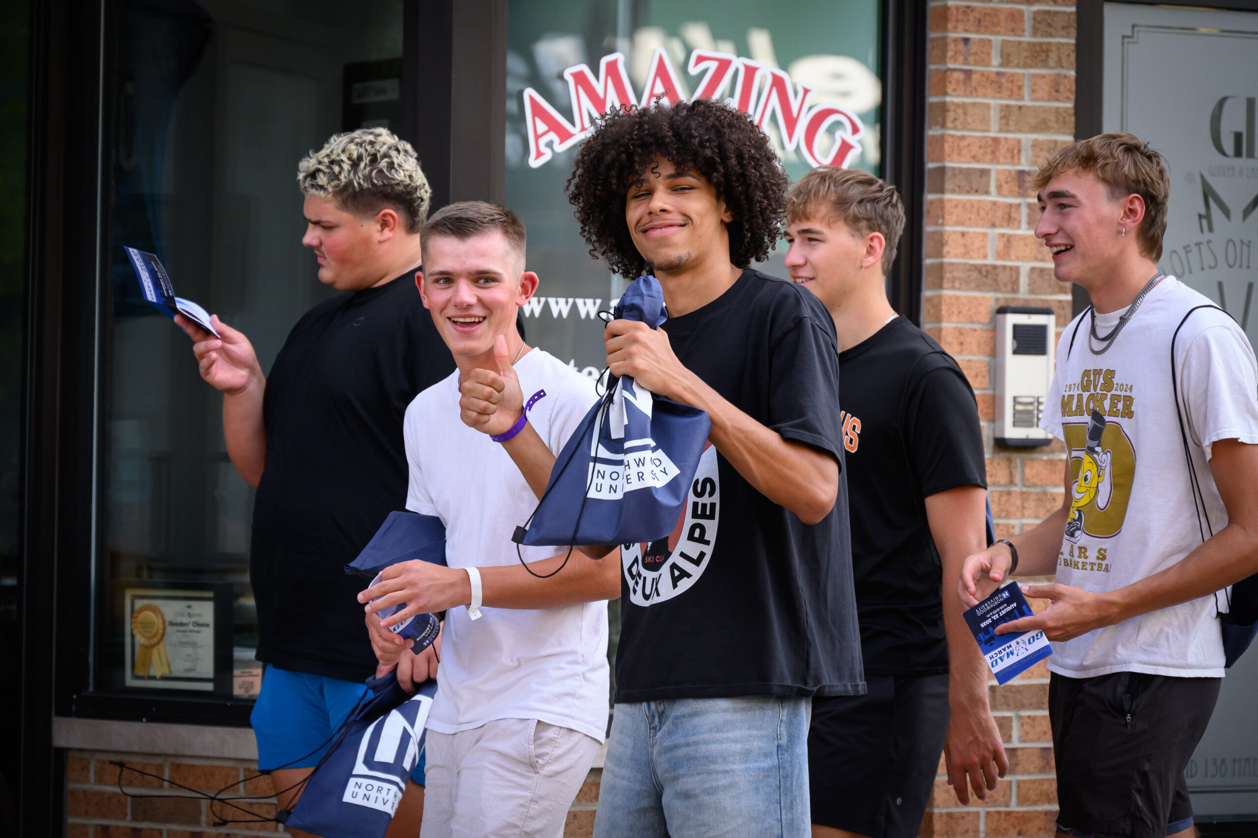 Five young men stand and walk outside a storefront, holding pamphlets and smiling, with one giving a thumbs up and another holding a microphone.