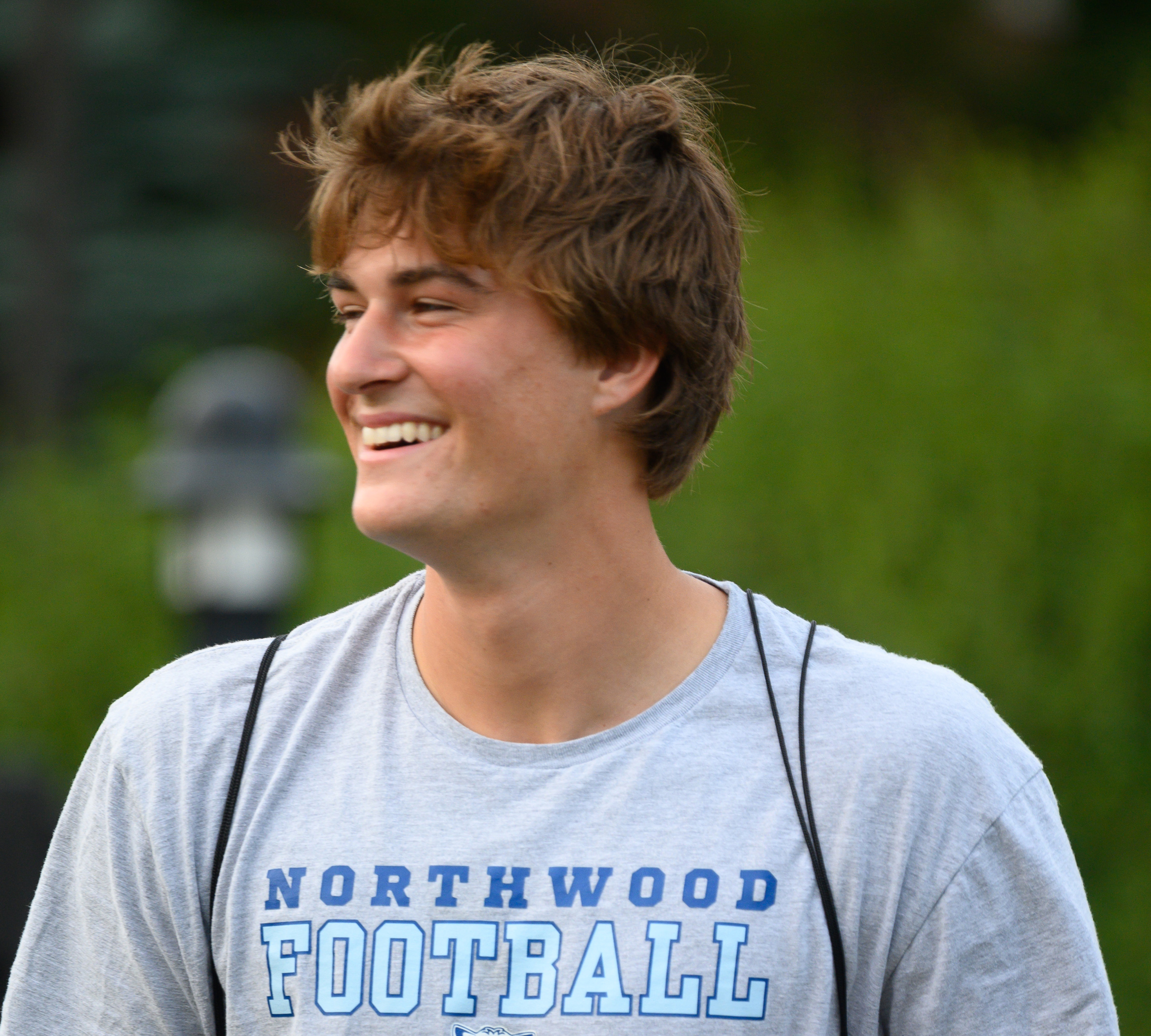 Young man smiling outdoors, wearing a gray "Northwood Football" T-shirt, with greenery visible in the blurred background.