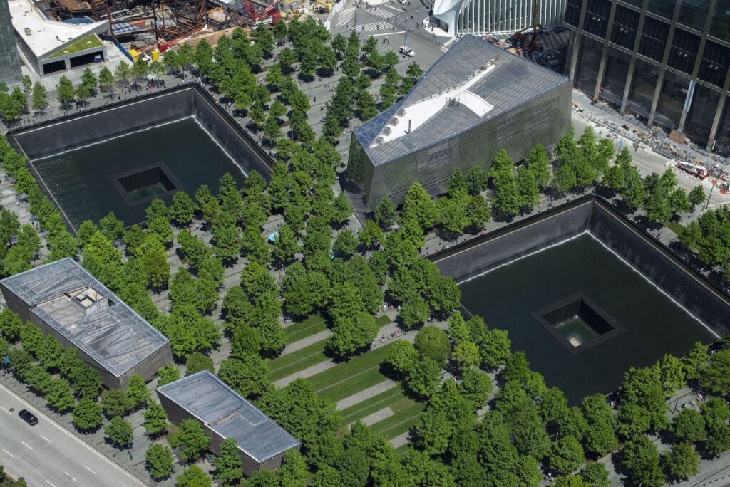 Aerial view of the 9/11 Memorial in New York City, showing two large square reflecting pools surrounded by green trees and nearby modern buildings.