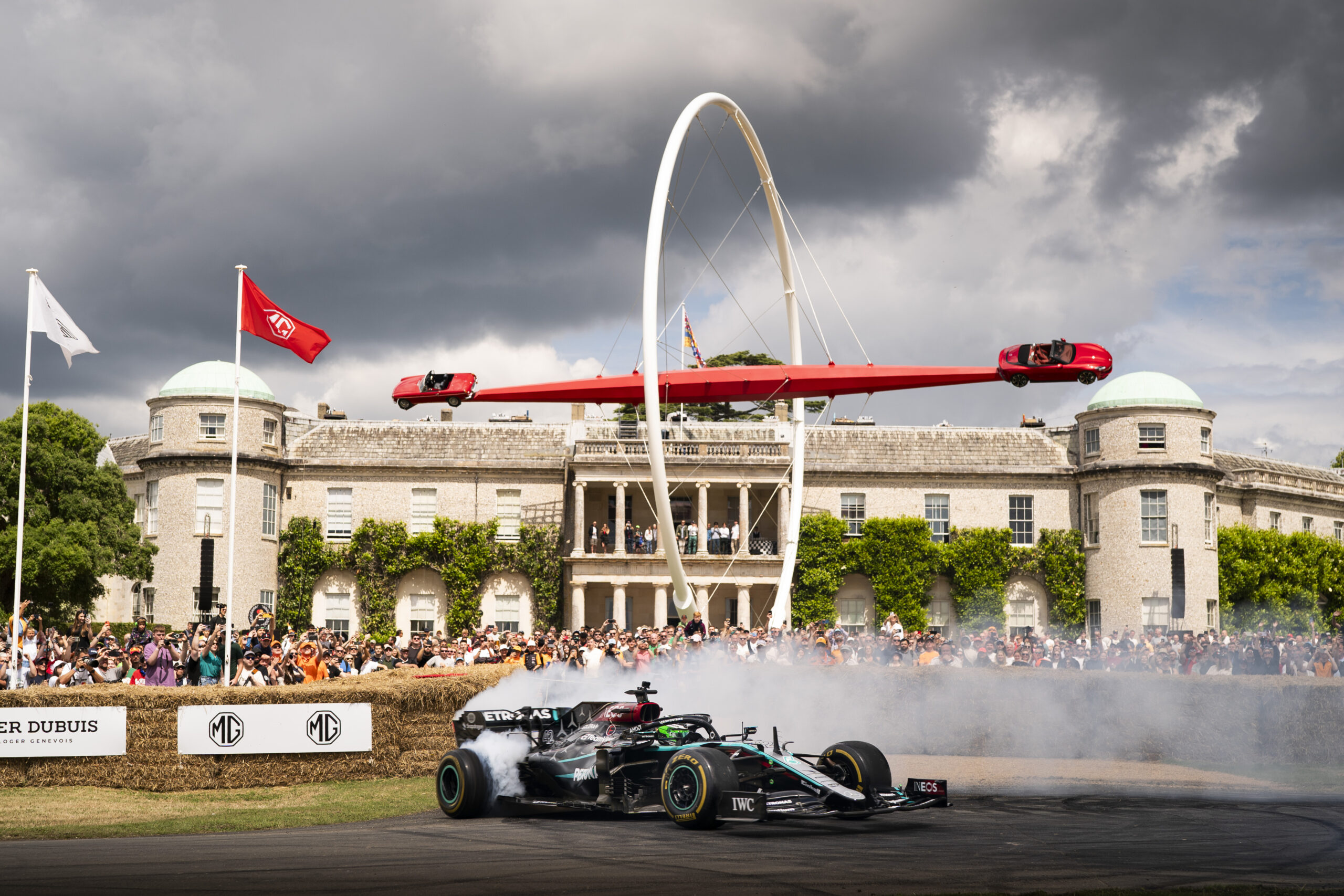 A Formula 1 car drifts and creates smoke in front of a large crowd and a historic building at Goodwood Road Racing Club, with a race car sculpture displayed on an arch overhead.