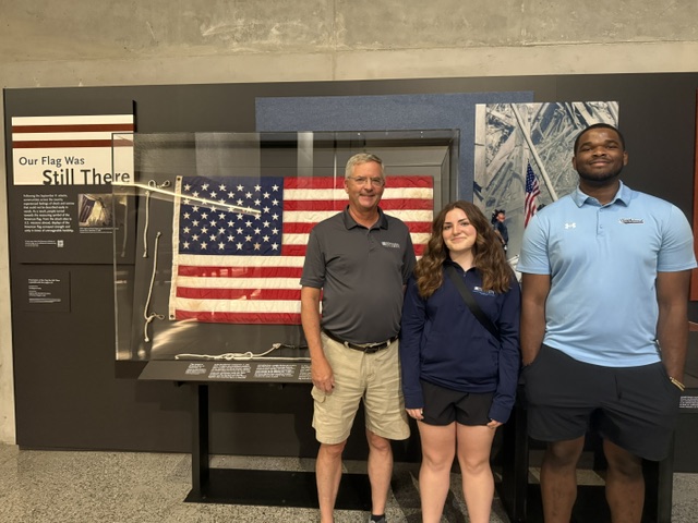 Three people stand in front of a display case containing a U.S. flag at a museum exhibit titled "Our Flag Was Still There.