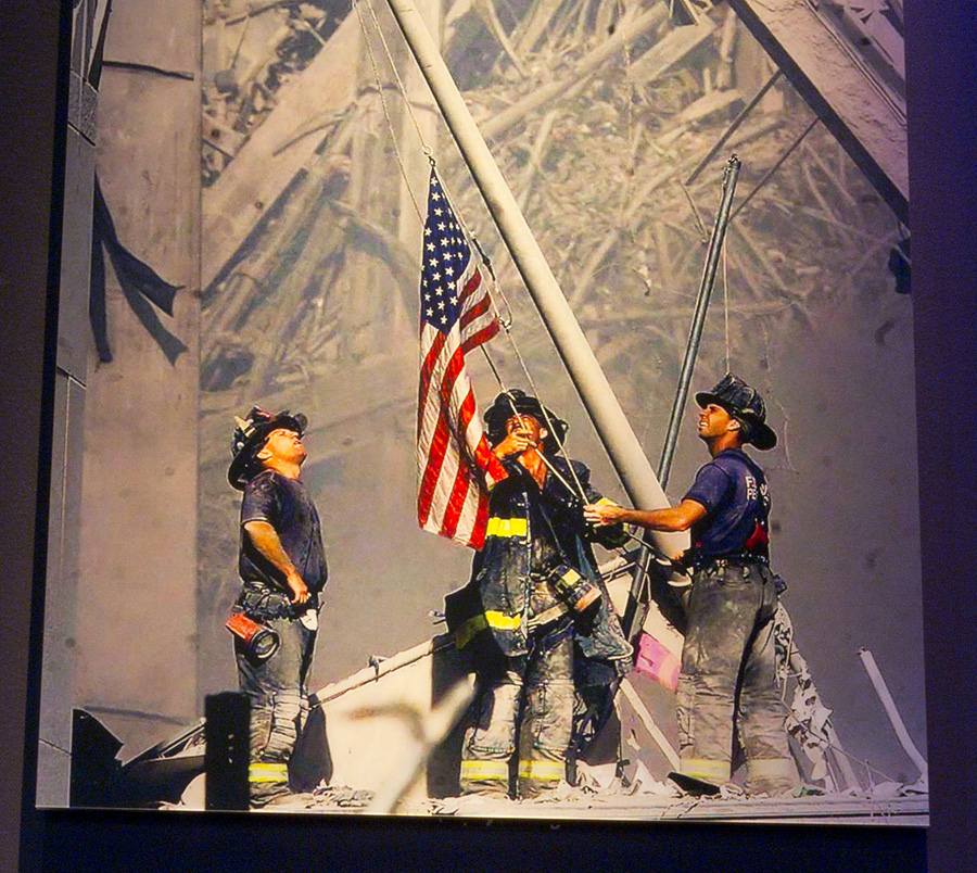 Three firefighters raise an American flag amidst rubble and debris from a collapsed structure in daylight.