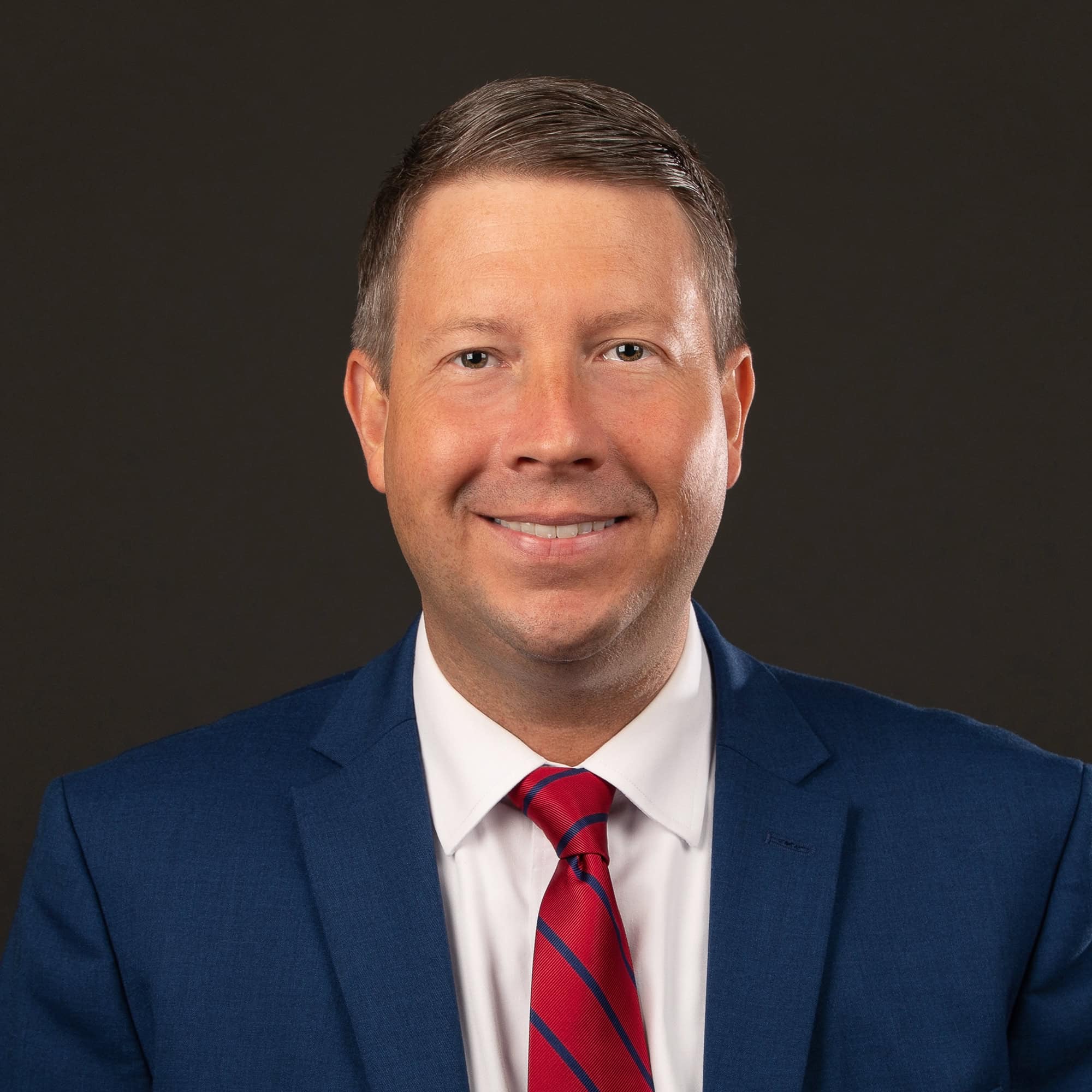 Man in a blue suit, white shirt, and red striped tie smiles at the camera against a plain dark background, presenting an approachable appearance perfect for providing information for parents.