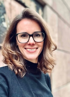 Woman with shoulder-length brown hair and glasses smiling, wearing a black top, standing in front of a blurred outdoor stone wall background—ready to share helpful information for parents.