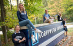 Five students pose on and around a large Northwood University sign outdoors, surrounded by trees with autumn leaves.