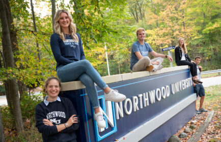 Five students pose on and around a large Northwood University sign outdoors, surrounded by trees with autumn leaves.