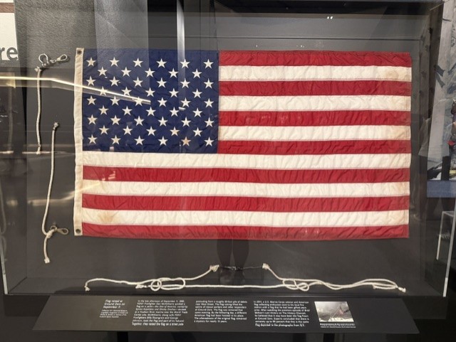 An American flag displayed in a glass case with informational plaques below and white ropes attached to the flag’s grommets.