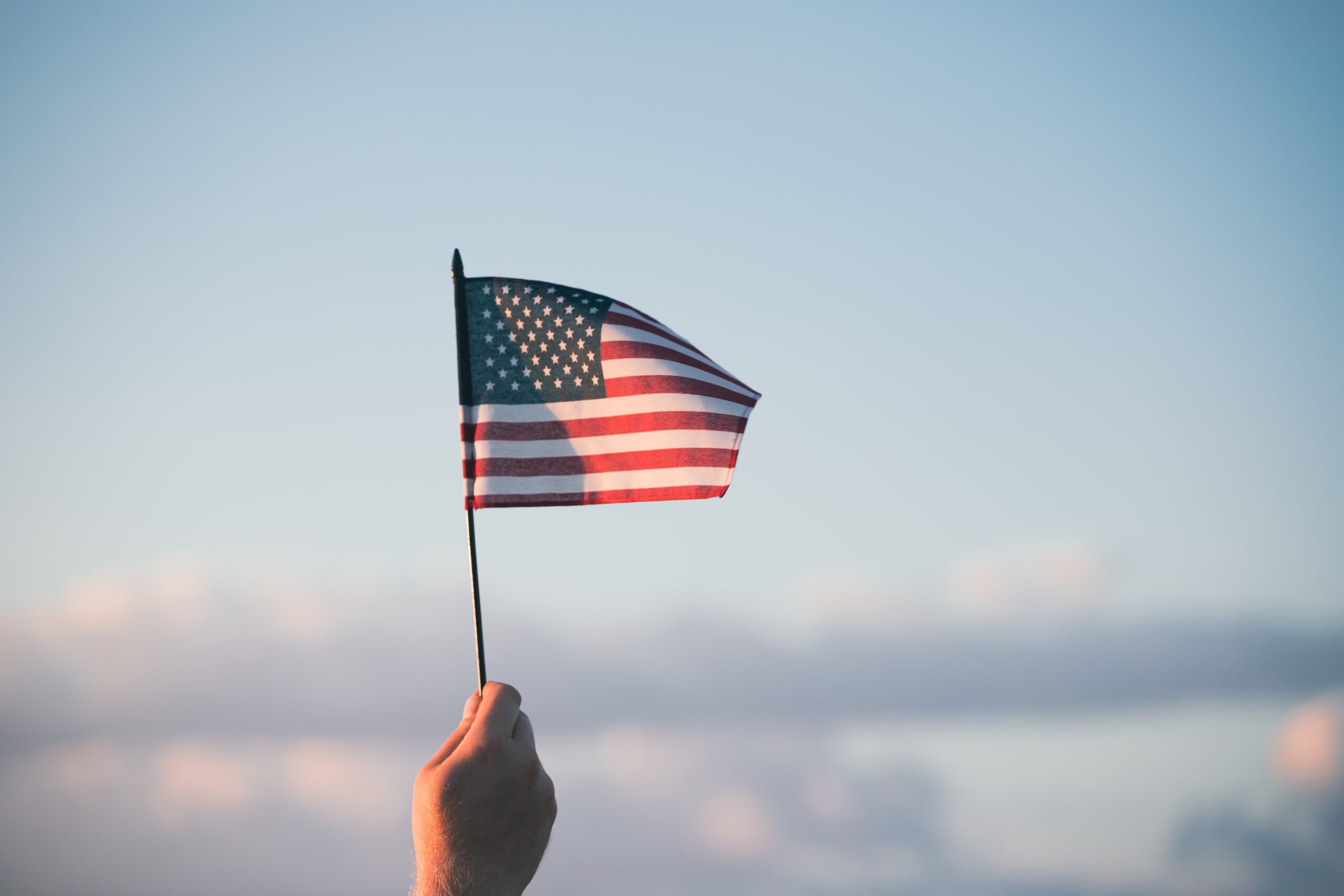 A hand holds a small American flag against a blue sky with soft clouds in the background.