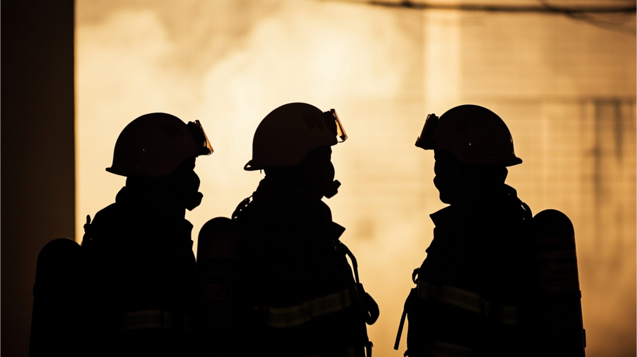 Three firefighters in full gear stand in silhouette against a smoky, backlit background.