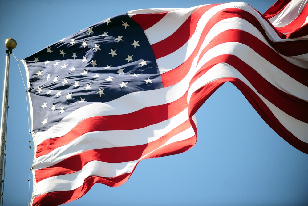 The United States flag waves in the wind against a clear blue sky, showing its stars and stripes in red, white, and blue.