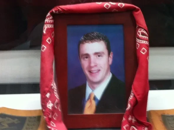 A framed portrait of a smiling man in a suit and tie, draped with a red bandana, displayed between two wooden plaques in honor of community recognition on the 25th anniversary.