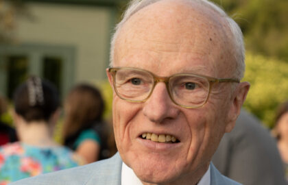 Older man wearing glasses and a light blue suit smiles outdoors at the Freedom Seminar hosted by Northwood University, with people and greenery blurred in the background, evoking the presence of an esteemed American Historian.