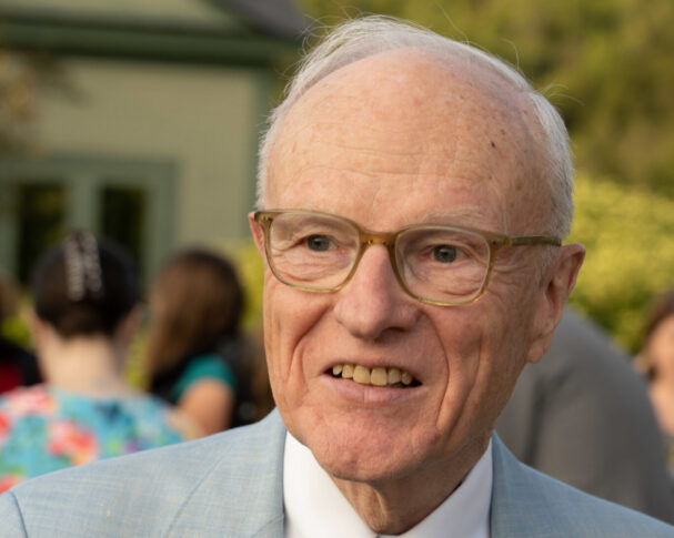 Older man wearing glasses and a light blue suit smiles outdoors at the Freedom Seminar hosted by Northwood University, with people and greenery blurred in the background, evoking the presence of an esteemed American Historian.
