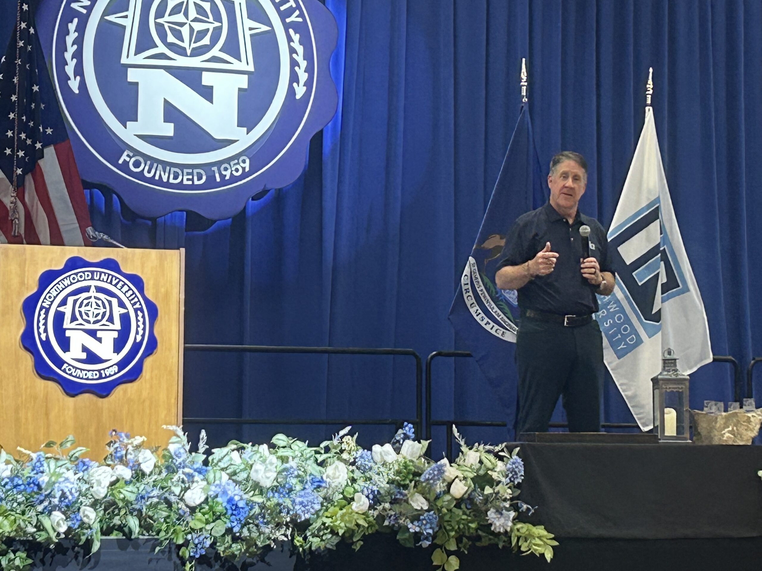 A man speaks on stage at a podium with Northwood University branding, highlighting the Economic Impact of Northwood in Michigan, with blue curtains and floral arrangements in the foreground.