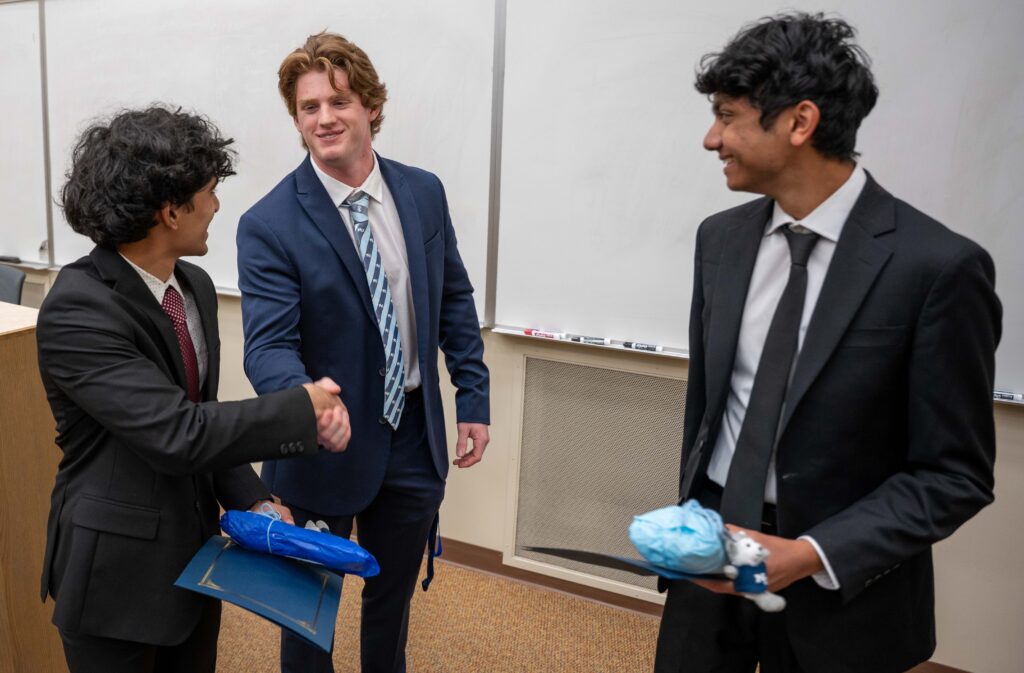 Three men in suits stand in a classroom; two are shaking hands while the third smiles and holds a blue-wrapped object.