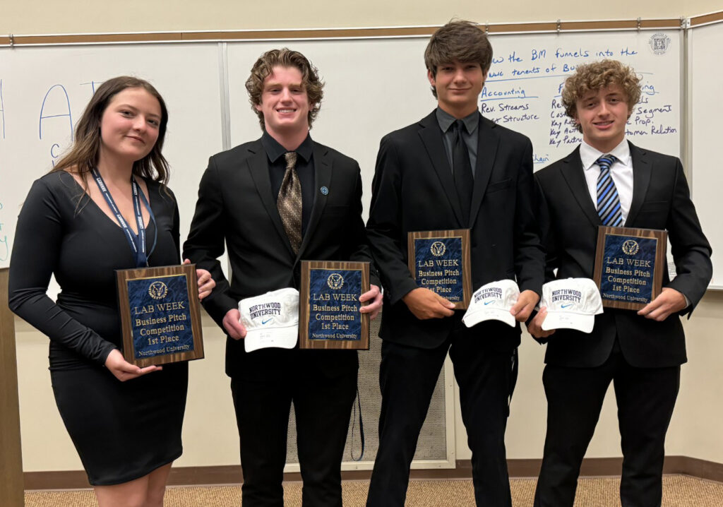 Four young adults in formal attire stand indoors holding plaques and baseball caps, posing for a group photo in front of a whiteboard covered in handwritten notes.