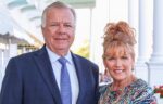 A man in a blue suit and tie stands next to a woman in a colorful dress with her hair up; they are both smiling outdoors near white columns, celebrating the Naegele Award at Northwood University.