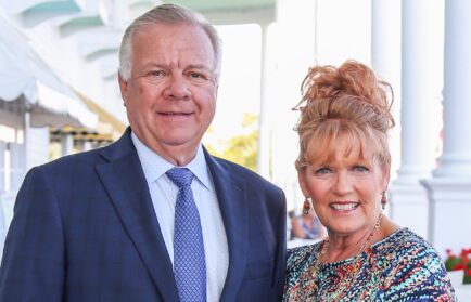 A man in a blue suit and tie stands next to a woman in a colorful dress with her hair up; they are both smiling outdoors near white columns, celebrating the Naegele Award at Northwood University.