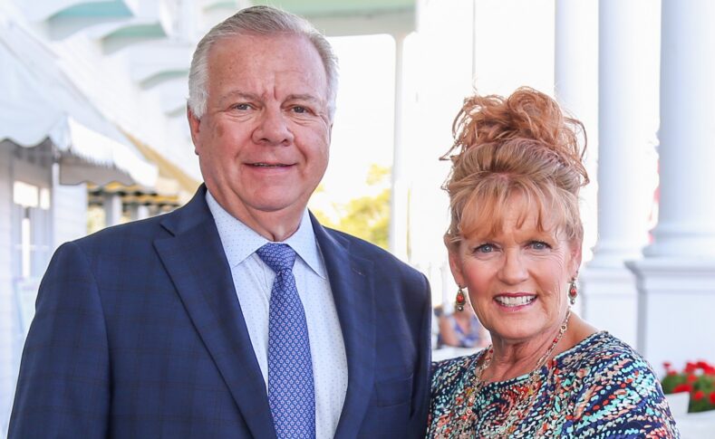 A man in a blue suit and tie stands next to a woman in a colorful dress with her hair up; they are both smiling outdoors near white columns, celebrating the Naegele Award at Northwood University.