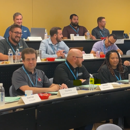 A group of people sit in a classroom setting at the university with laptops, drinks, and papers on desks, attending what appears to be a professional workshop or meeting focused on automotive topics.