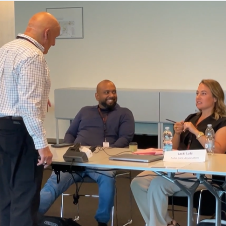 Three people are in a university meeting room; one man stands while two others sit at a table with nameplates, water bottles, and papers displaying automotive project details.