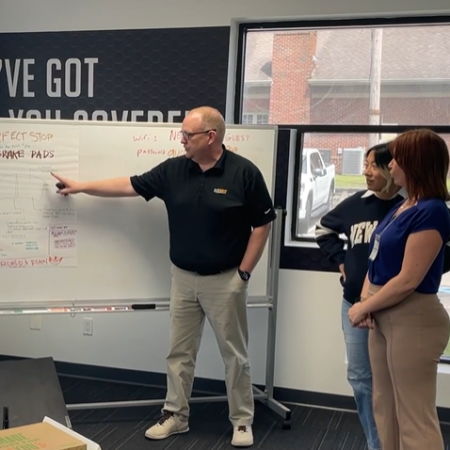A man points at a whiteboard with notes while two women stand nearby, listening. They are in a university office with a large window and another whiteboard in the background, discussing an automotive project.