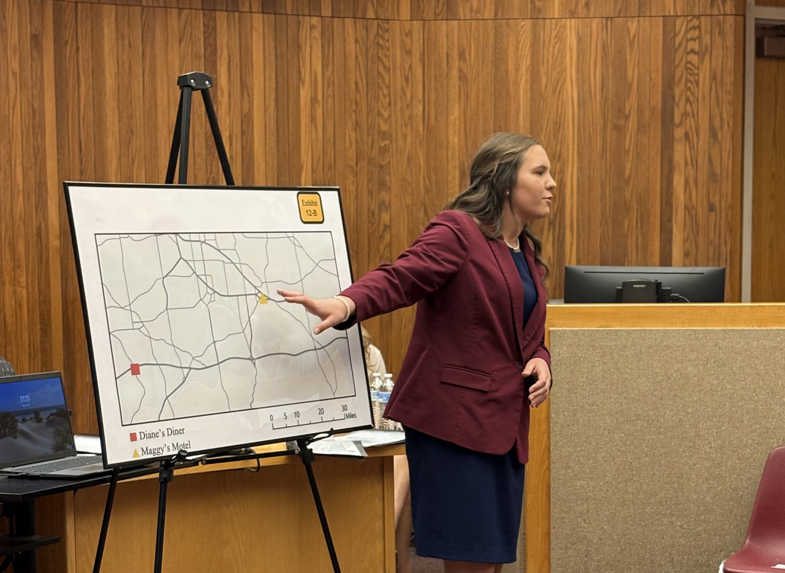 A woman in a maroon blazer points to a map on an easel in a wood-paneled courtroom, presenting information to an unseen audience.