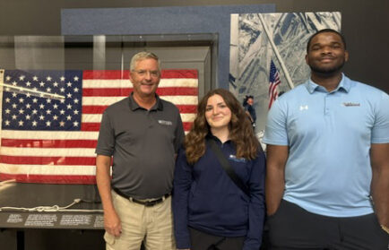 Three people stand in front of a framed American flag and wall display, posing and smiling for the photo.