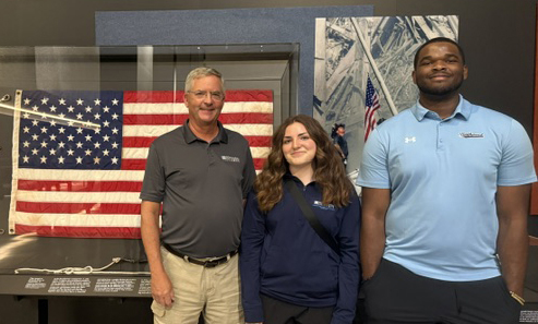 Three people stand in front of a framed American flag and wall display, posing and smiling for the photo.