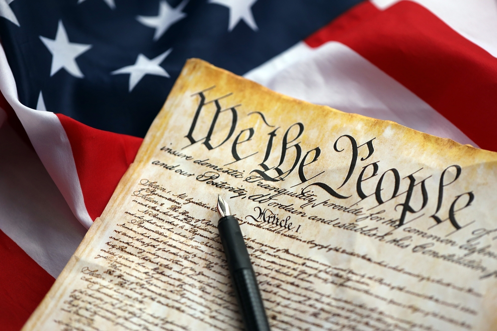 A close-up of the U.S. Constitution with the words โWe the People,โ a black pen, and a blurred American flag in the background, making it a fitting tribute for Constitution Day.
