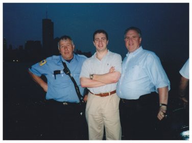 Three men posing together outdoors at dusk; one wears a light blue police uniform, and the other two are in casual shirts and pants—gathered for community recognition. The city skyline is faintly visible in the background.