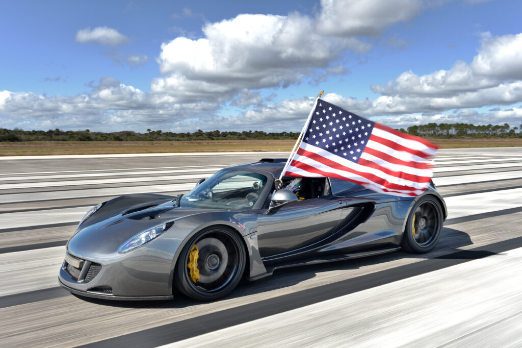A gray sports car drives on a runway while a driver waves a large American flag out the window under a partly cloudy sky.